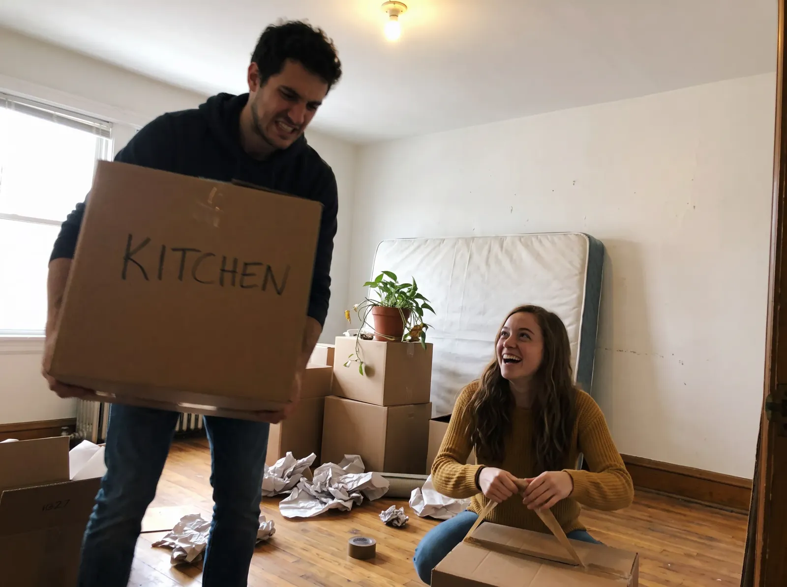 He carries a box marked KITCHEN while she unpacks on the floor