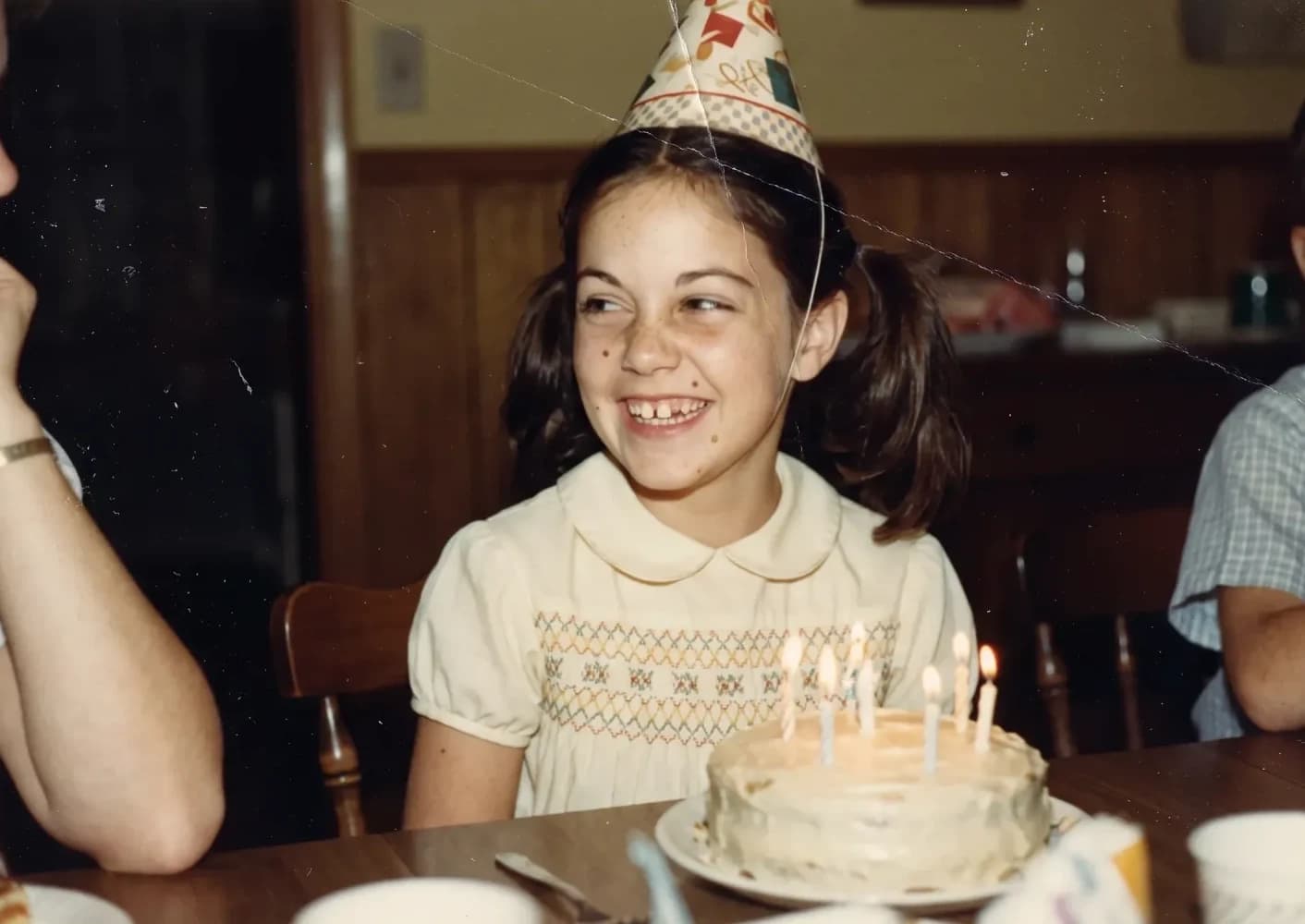 A girl in a party hat smiling behind her birthday cake