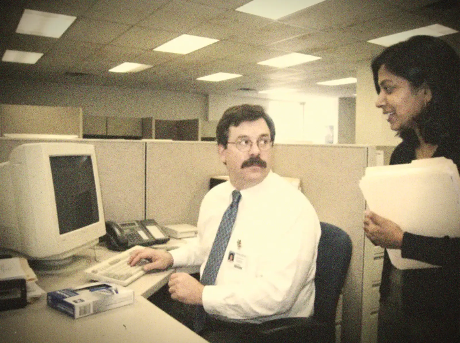 A young man in a tie at an office computer, talking with a colleague